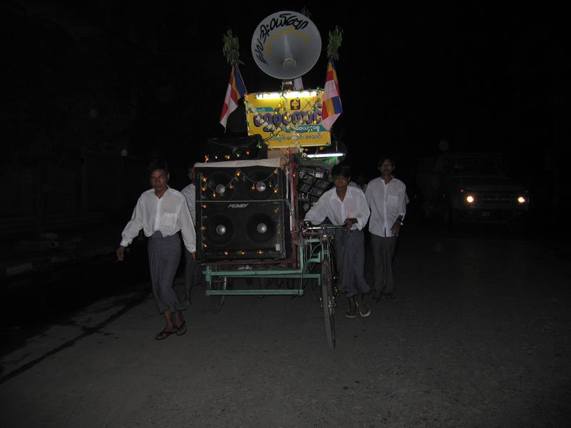 this is a travelling band with instruments hooked up to amps and speakers on wheels, wandering the streets at night and blaring music
