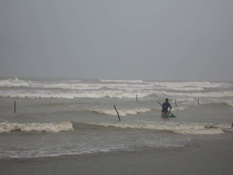 one lone fisherman standing close to the shore in the rough ocean and the heavy rain, still trying to catch something
